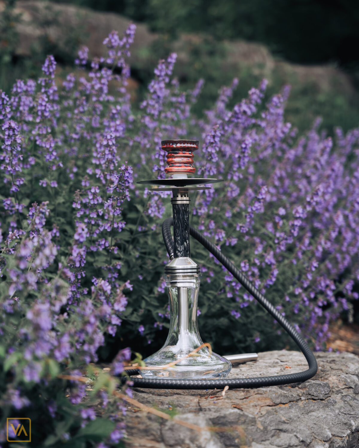 Steamulation Xpansion Mini Posed on rock surrounded by flowers and greenery with a Mason Helyx Bowl, Konus 1 Handle on Carbon Dschinn Silicone and a AO Stainless Steel Support Strainer